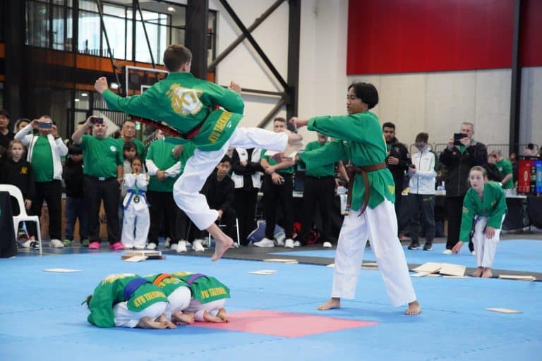 A taekwondo South Windsor demonstration shows a boy in a green belt uniform jumping over two crouched teammates whilst kicking a board held by another student, with an audience watching in the background.